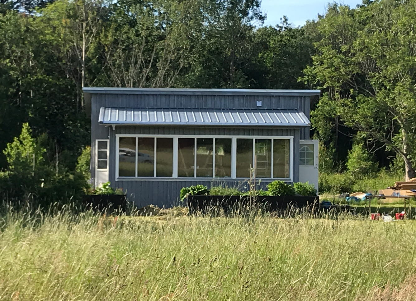 Ett litet grått hus som står bakom en äng före ett skogsparti. Fönsterpartier vetter ut mot ängen.