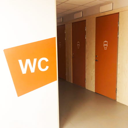 Toalettdörrar med en toalettsymbol. På väggen framför står WC på en orange färgplatta.