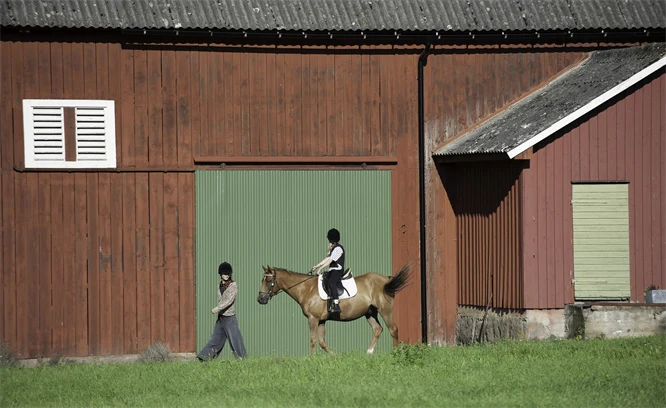 Flicka rider på en häst vi en röd ladugård. Person går bredvid hästen.