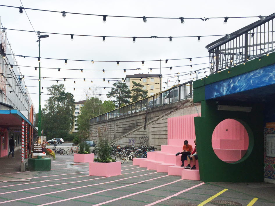 Bilden visar tillfälliga gatumöbler och blomsterlådor i klara färger i gult och rosa samt ett tak av ljusslingor markerar platsen mellan pendelstågsstationen och gångtunneln som leder till Poseidons torg.