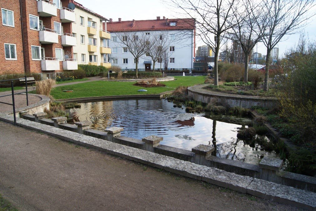 Tre bostadshus i olika färger med en vattenfylld damm framför.