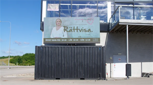 Foto på en stor bildskärm med reklam för Kvinnojouren, ståendes på en svart container vid en affär.