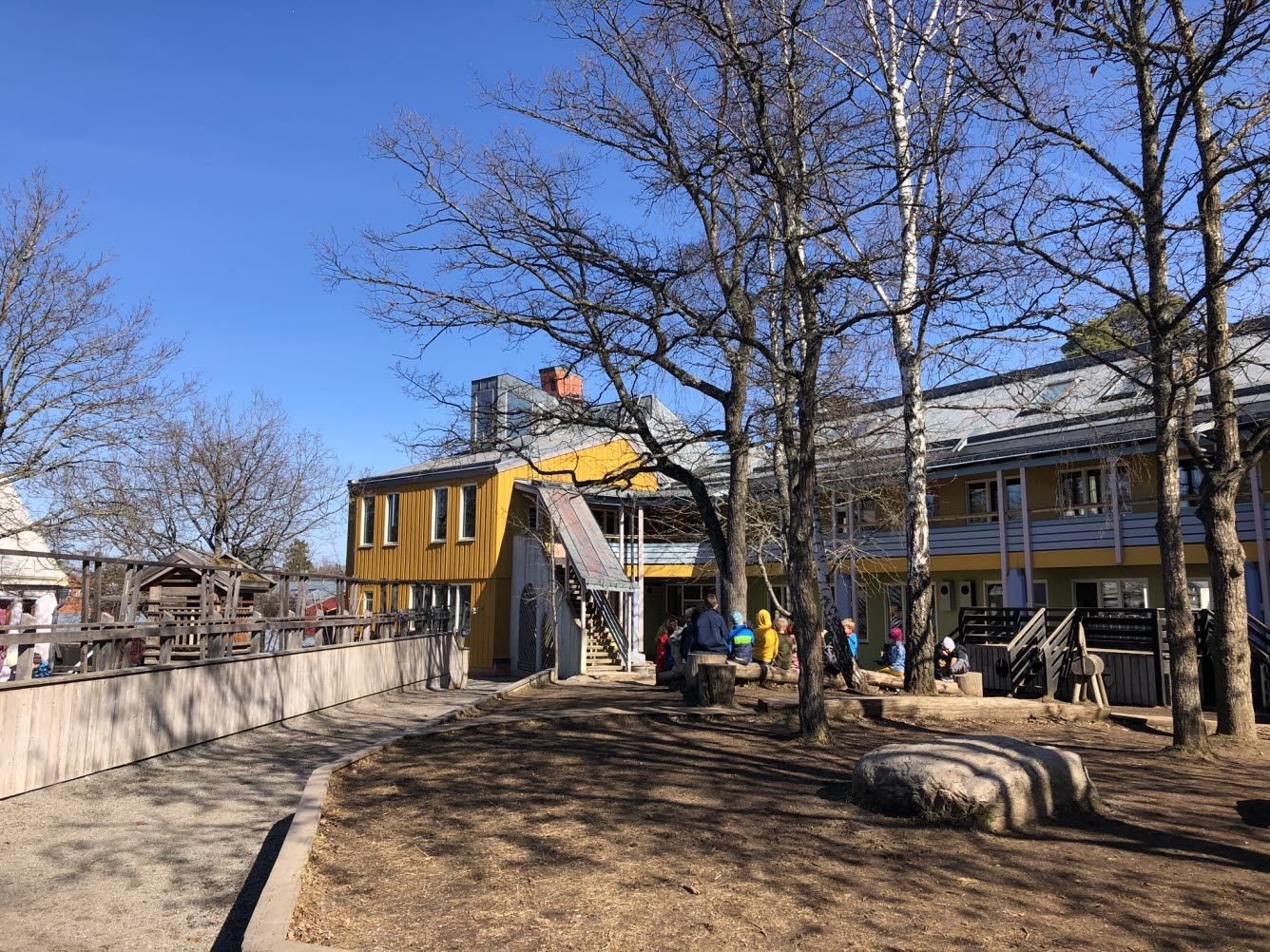 Skolbyggnad i bakgrunden med gräsmatta och stora träd framför.