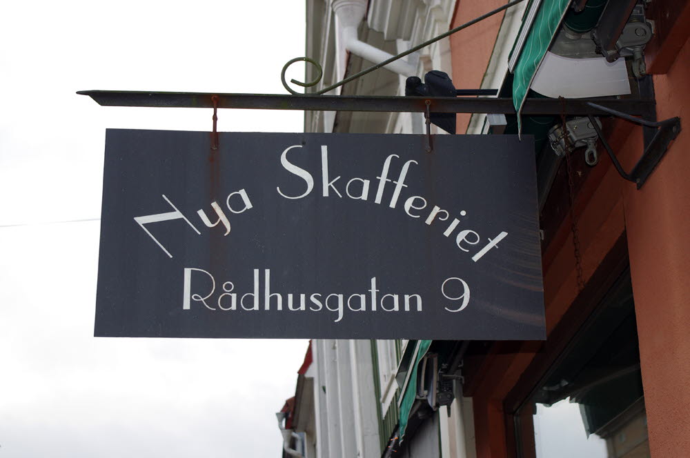 Foto på svart pendelskylt av metall med texten Nya Skafferiet Rådhusgatan 9 hängandes ut från en husfasad..