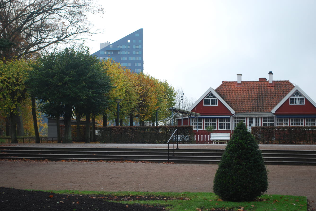 Bilden föreställer en parkmiljö med träd, planteringar och grusgångar samt en äldre träbyggnad.