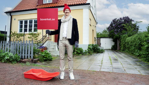Man med mössa och halsduk står med en röd pulka framför ett gult hus. Det är barmark.