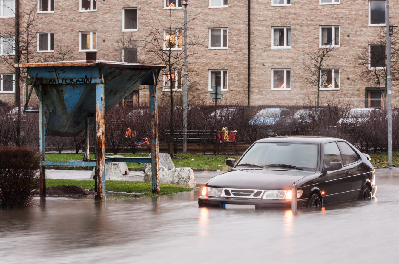 Bil kör på översvämmad gata i bostadsområde med flerfamiljshus.