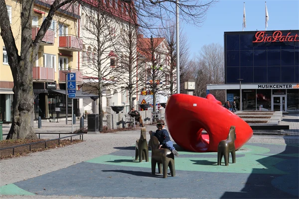 Lekplats på ett torg. Träd och byggnader runt torget. En pojke leker på lekplatsen.