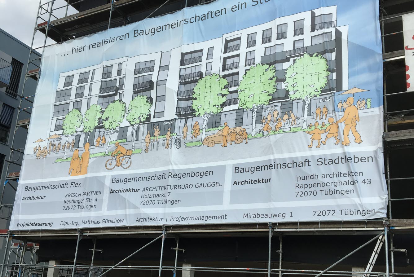 En idéskiss på bostadsbygge hänger på en byggställning framför bostadsbygge. Texten på affischen är på tyska.