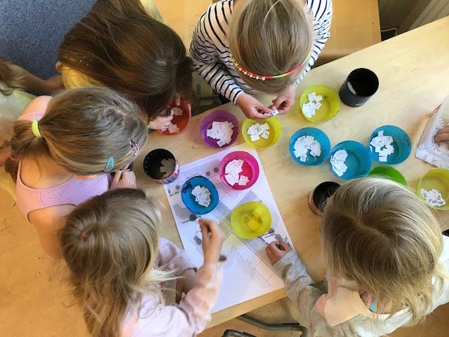 Foto på barn runt ett bord som har vita lappar i färgglada skålar framför sig.