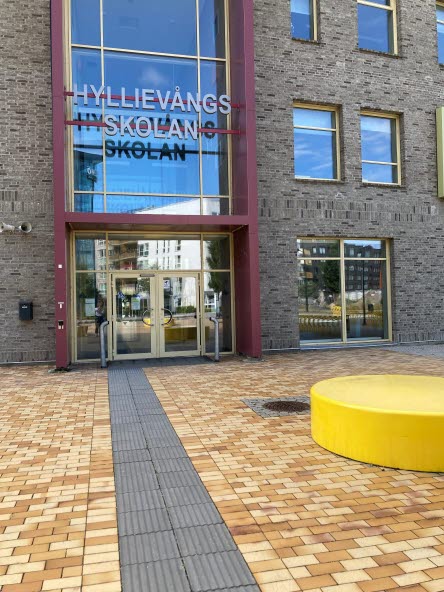 Fasad på Hyllievångsskolan. Marken framför skolan är lagd av orange marktegel. Svarta plattor skapar en gång in i byggnaden.