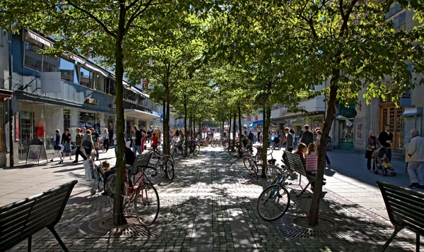 Foto av en gågata med träd som ger skugga, folk i rörelse.