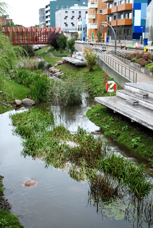 Grönområde utmed slingrande bäck i stadsbebyggelse. Sluttningar med gräs och buskar.