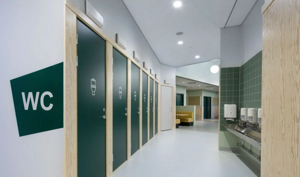 Gröna toalettdörrar med en toalettsymbol på dörren. Utanför står WC med stora bokstäver på väggen. Till höger finns handfat.