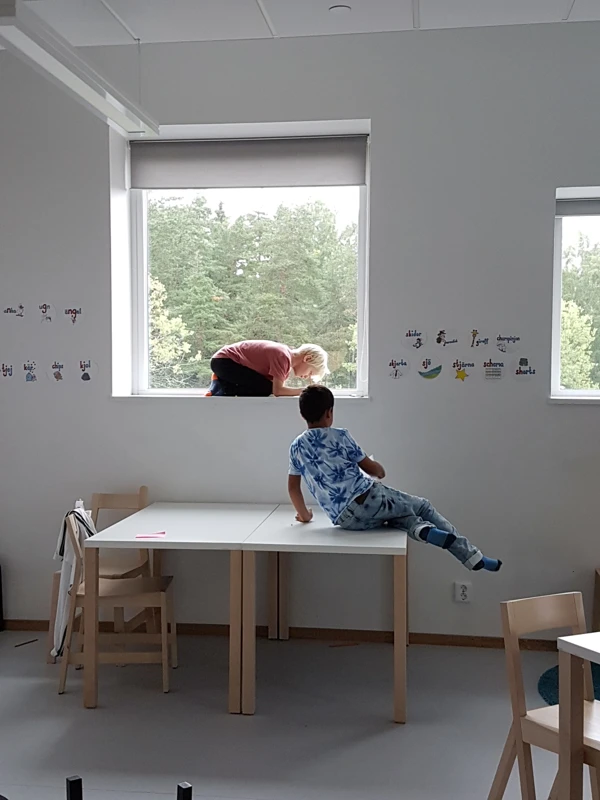 Två barn i ett klassrum. Det ena barnet sitter på ett bord och det andra är hopkrupet i fönsternischen.
