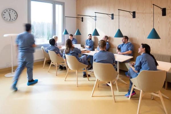 Blåklädd vårdperson på väg till ett konferensbord där flera andra blåklädda vårdpersoner sitter.