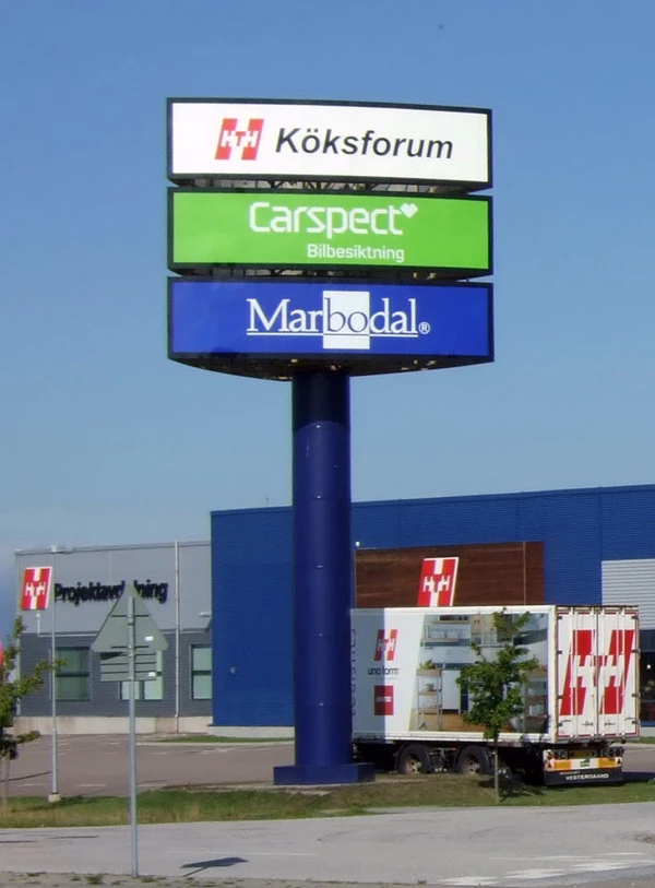 Foto av en hög stolpe med tre skyltar med texten Köksforum, Carspect Bilbesiktning och , Marbodal vid ett köpcentrum.